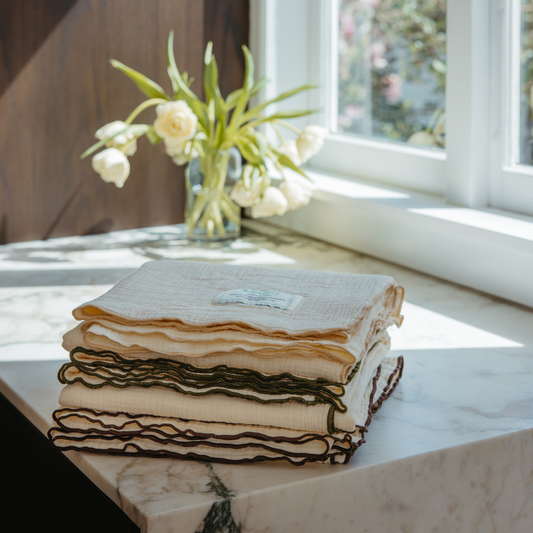 A stack of folded cotton muslin cloths with ruffled edges, some in cream and some with dark olive green trim, displayed on a white marble table next to a window with white tulips in the background.
