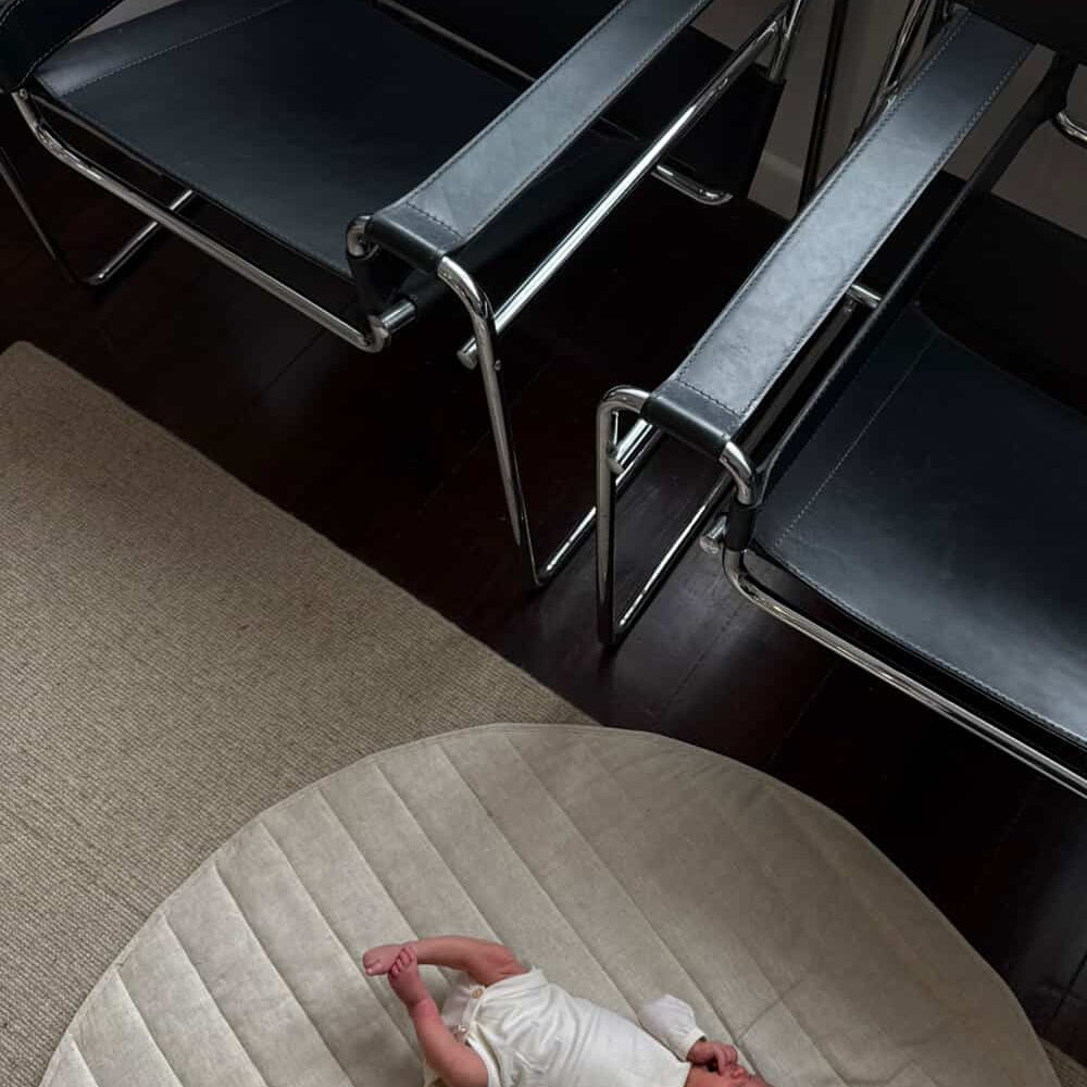 Newborn baby lying on a round cushion in an office setting with chairs.